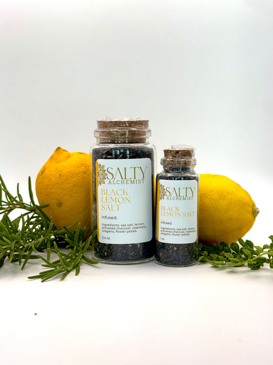 infused gourmet salt for cooking and cocktail rims. black lava salt with lemon. Two glass jars of Salty for Alchemist Black Lemon Salt, one large and one small, styled with fresh lemons, rosemary and oregano . 